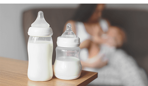 Two bottles of milk sitting on surface near woman breastfeeding her infant in background