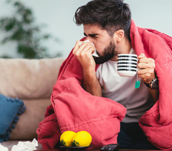 Sick person blowing their noose with a blanket over them sitting on a couch