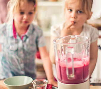 Two children helping make a pink smoothie in a blender at a kitchen counter