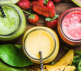 Birds-eye view of three different fruit smoothies in glass cups surrounded by a variety of fresh fruit