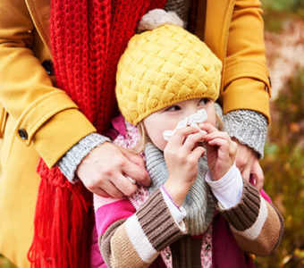 Child blowing their nose with a facial tissue while standing outside in front of an adult