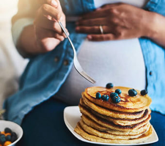 Pregnant person eating a stack of blueberry pancakes with their hand on their stomach