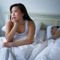 Person sitting up in bed, looking to the side thoughtfully, while other person is asleep